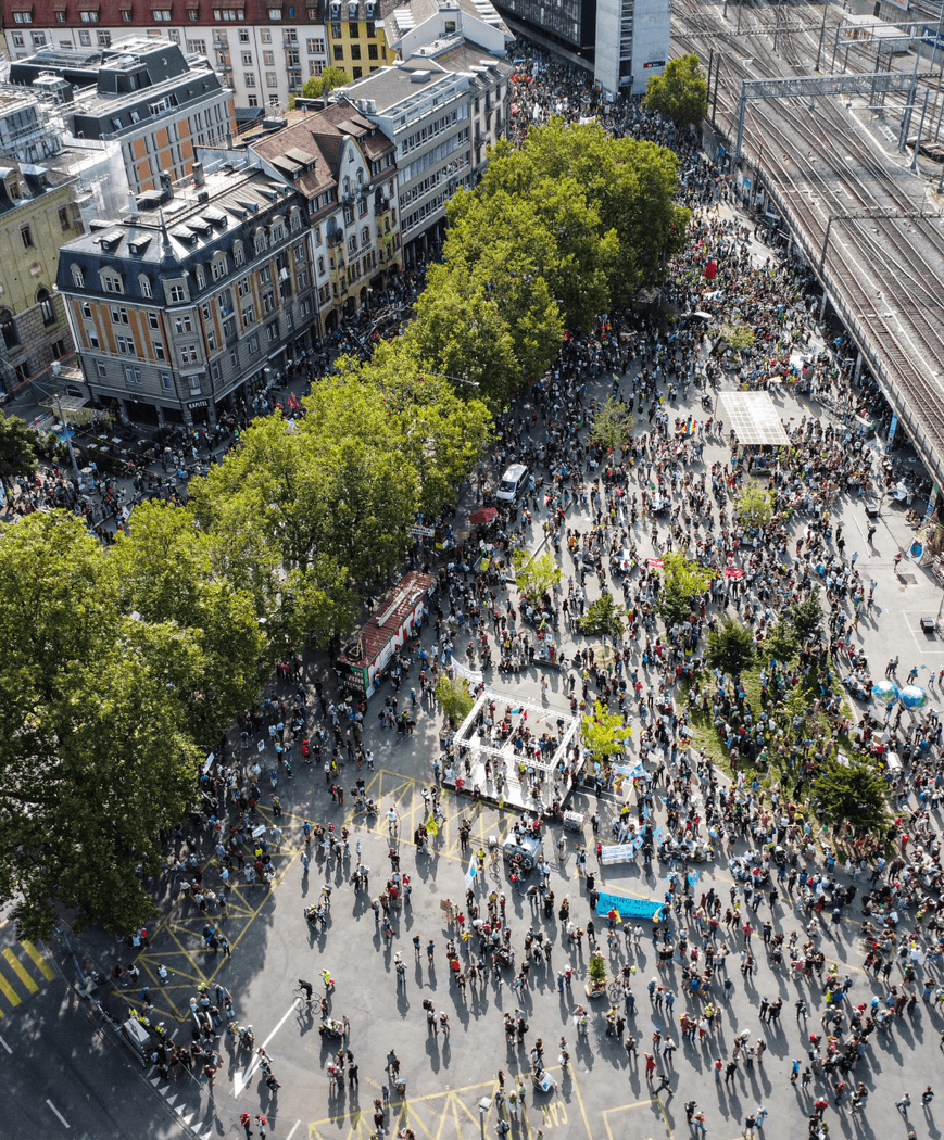 Eine Stadt aus der Vogelperspektive. Es versammeln sich viele Leute unter Bäumen und um Häuser um eine Bühne.